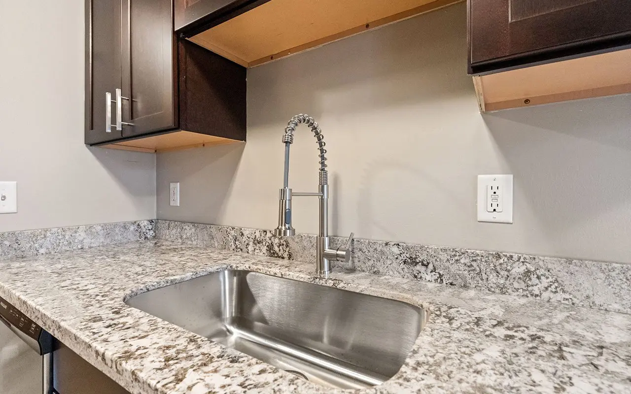 Modern kitchen counter with a stainless steel sink, high-arc faucet, granite countertop, and dark wood cabinets. Cabinet doors above are open or missing.