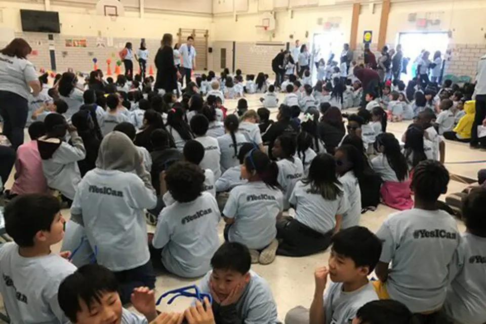 A large group of children wearing matching "#YesICan" shirts sit on a gym floor, facing the front, while adults stand and move around in the background.