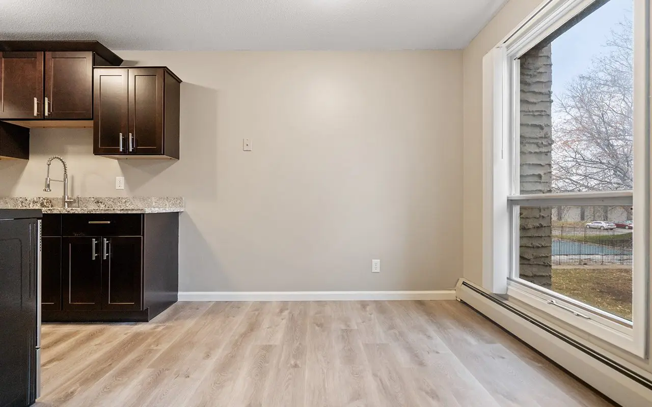 Unfurnished room with light wood flooring, beige walls, a large window, and a kitchen area with dark cabinets and a sink on the left side.