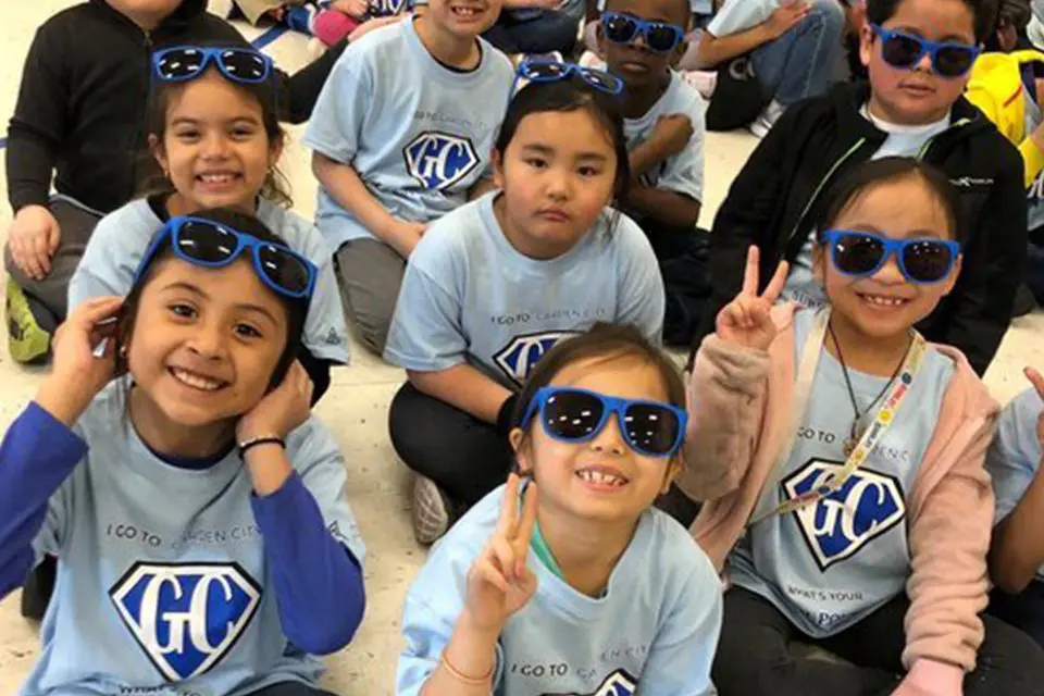 A group of young children wearing blue sunglasses and matching blue T-shirts sit together, some smiling and making peace signs.