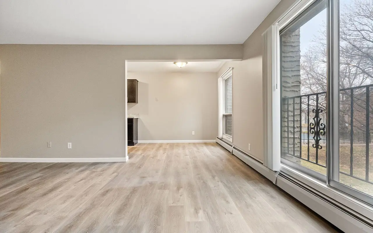Unfurnished living room with light wood flooring, beige walls, large windows, and a sliding glass door leading to a balcony with a view of trees.