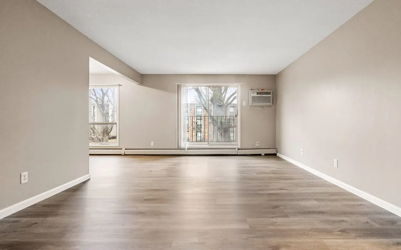 Empty living room with light brown wood flooring, beige walls, a large window, and an air conditioning unit mounted above the window.