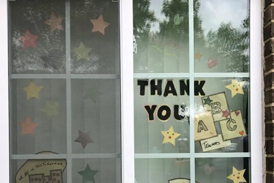 A window decorated with colorful paper stars and signs reading "THANK YOU" and "ABC Teachers," with reflections of trees visible on the glass.