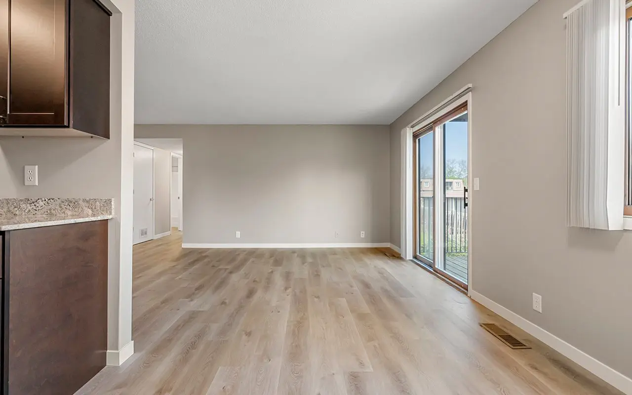 Empty living room with light wood flooring, beige walls, a sliding glass door leading to a balcony, and partial kitchen view on the left.