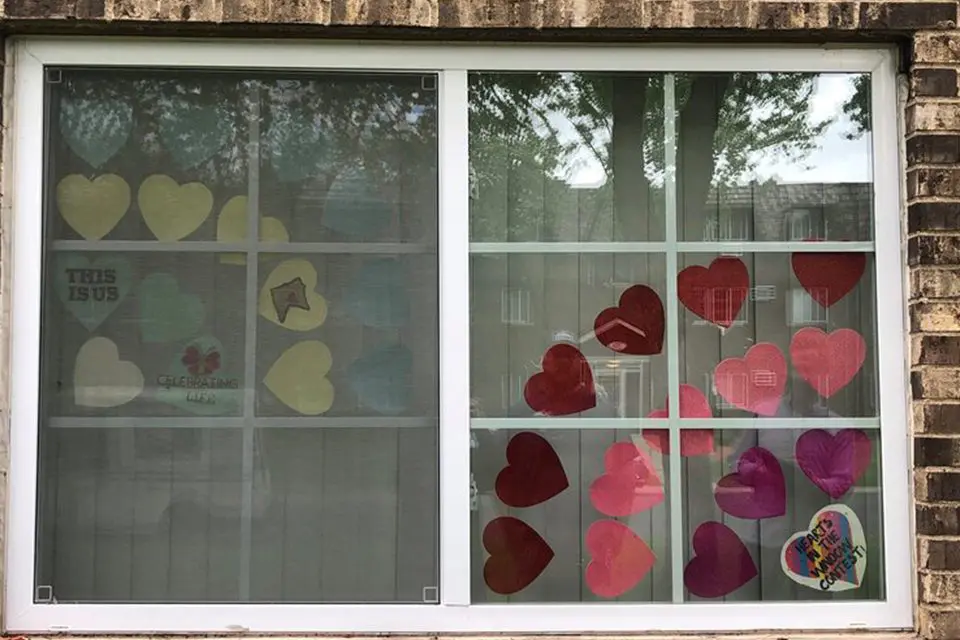 A window with paper hearts in various colors taped to the glass, some with messages written on them, and a view of trees and buildings reflected outside.