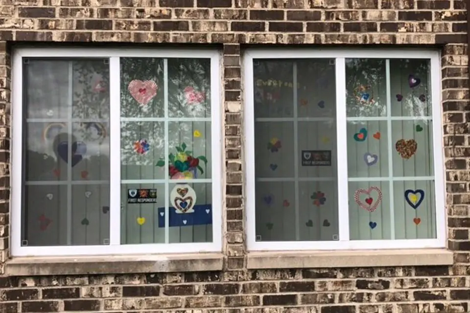 Two windows on a brick wall display paper cutouts of hearts, flowers, and rainbows, along with supportive signs taped to the glass.