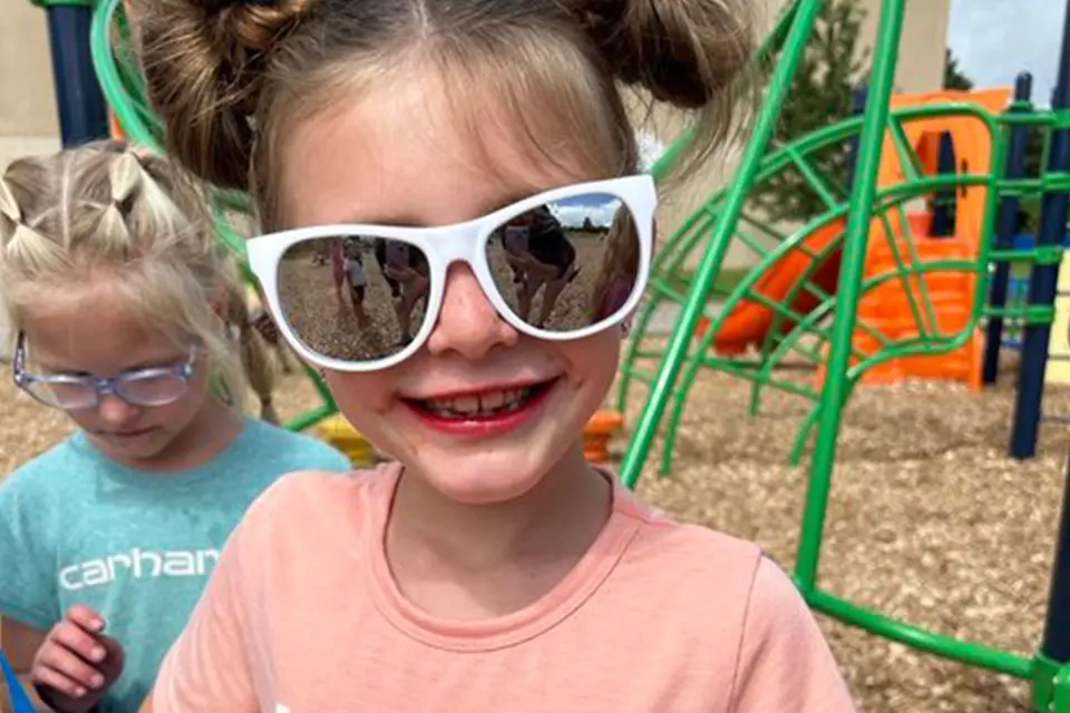 Two young girls on a playground; one in front wearing white sunglasses and a pink shirt, smiling, the other in the background with glasses and a blue shirt.
