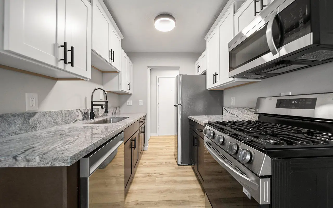 Modern galley kitchen with white cabinets, stainless steel appliances, marble countertops, and light wood flooring.
