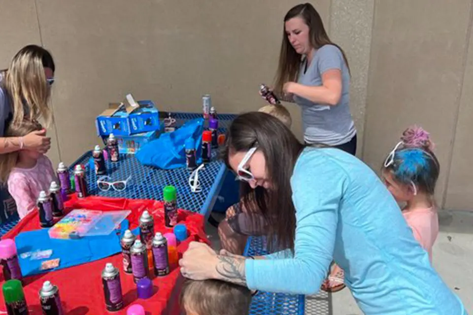 Adults and children gather at an outdoor table with spray cans, coloring the children’s hair and working on crafts.