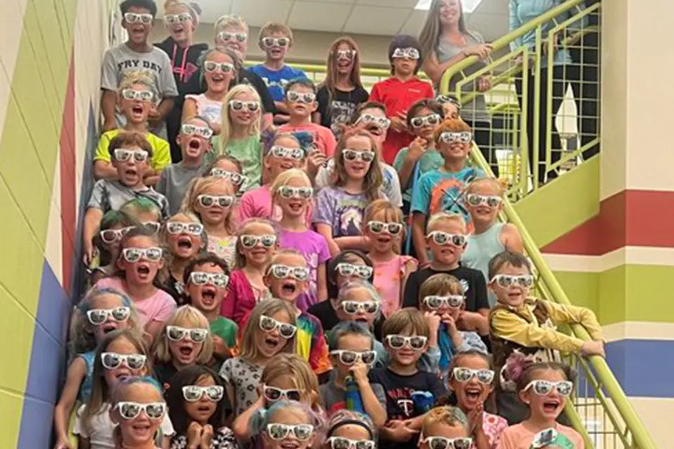 A large group of children wearing sunglasses sits and stands on a staircase indoors, smiling and looking at the camera; two adults stand at the top of the stairs.