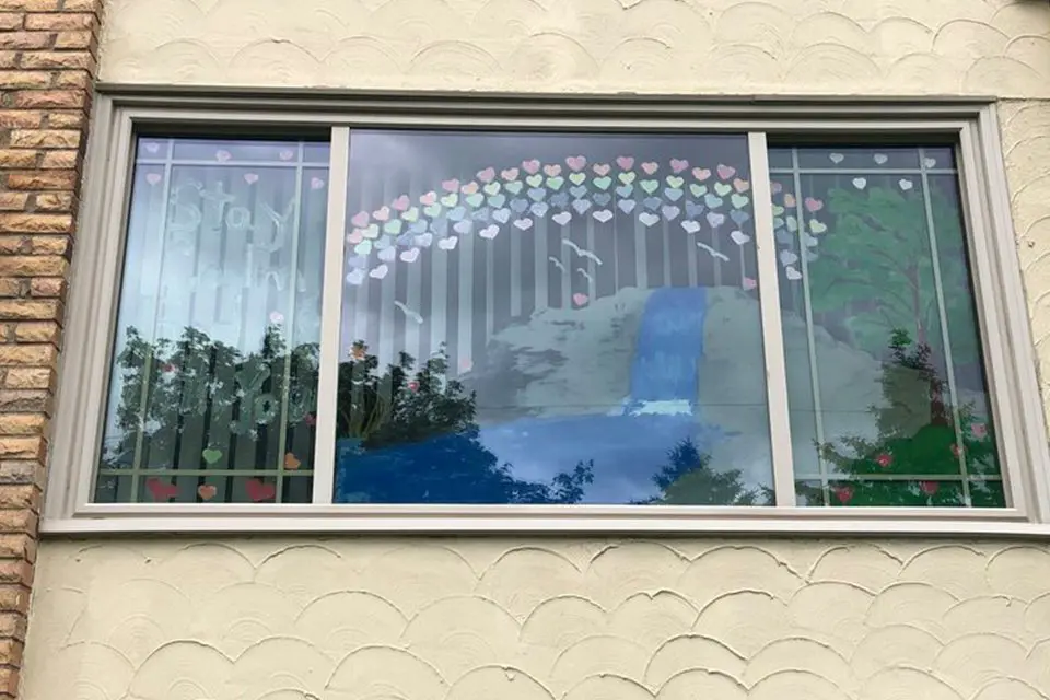 A large window displays a painted waterfall scene with trees, a rainbow made of paper hearts, and "Joy" spelled out on the left side. Some reflections of trees are visible on the glass.