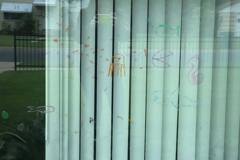 Vertical blinds with childlike drawings of sea creatures, including fish, a jellyfish, and a seahorse, made with colored markers on the window.
