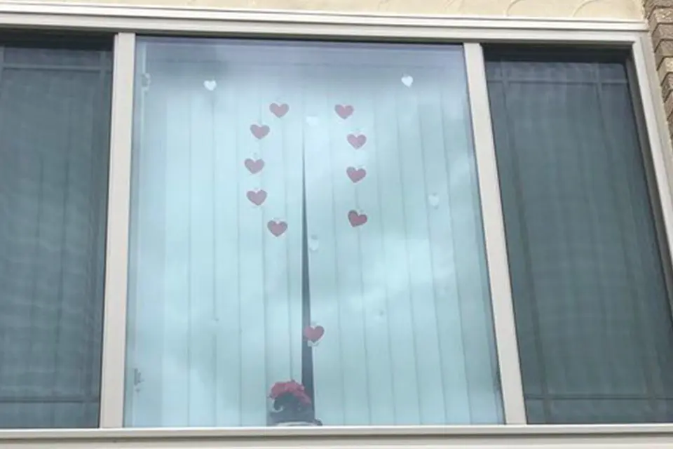 A window with vertical blinds is decorated with small red heart stickers arranged in the shape of the letter "U.