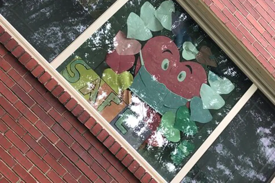 A large, colorful paper decoration of a smiling apple with hearts and the letters "S" and "L" is taped to the inside of a brick building window.