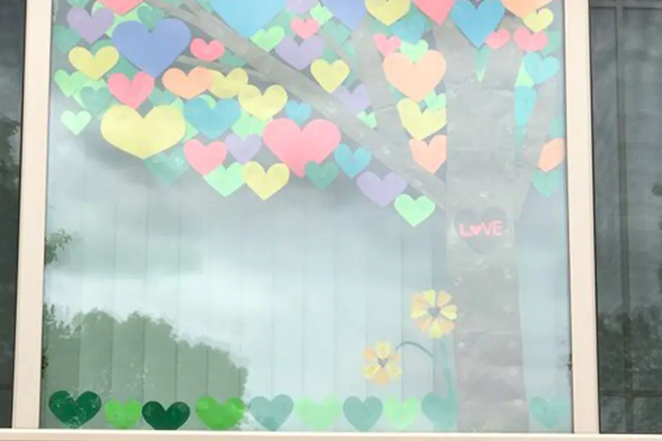 A window decorated with colorful paper hearts arranged as a tree, with additional heart and flower shapes along the bottom. The word "LOVE" is written on one heart.