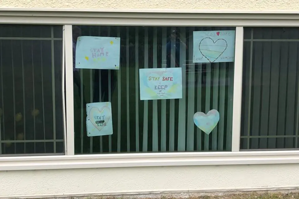 A window with five handmade signs, including messages like "Stay Home" and "Stay Safe," along with hand-drawn hearts and stars, displayed behind vertical blinds.