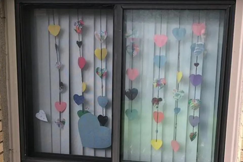 A window with vertical strings of colorful paper hearts hanging in front of white blinds.