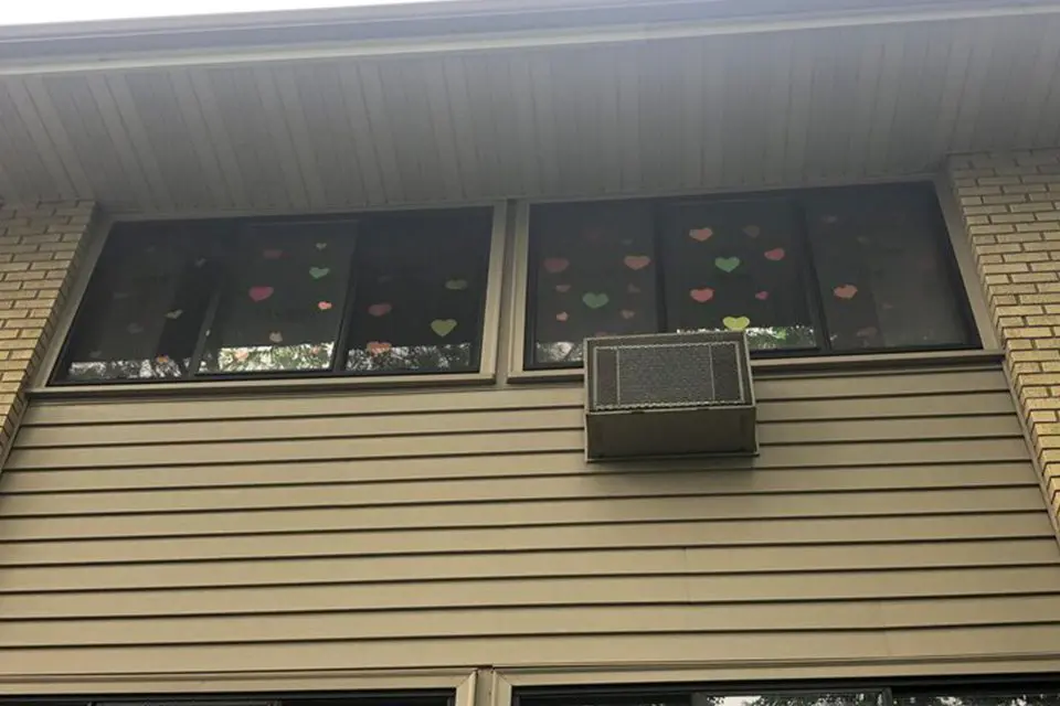 A view of an upper floor window decorated with colorful paper hearts; an air conditioner unit is installed in the center window.