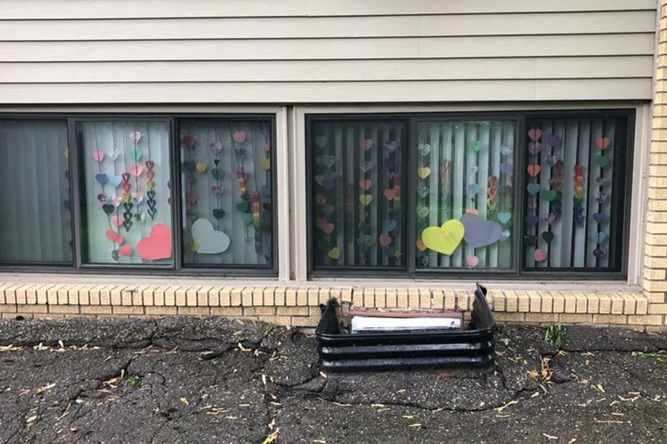 Colorful paper hearts are taped to the inside of two lower-level windows of a brick building, with a small planter in front on cracked pavement.