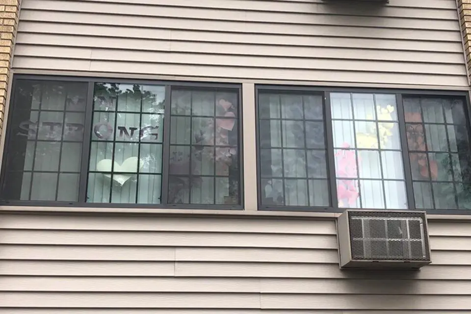 A window with paper cutouts spelling "STRONG," a heart shape, and colorful decorations taped to the glass; an air conditioning unit is installed below the window.