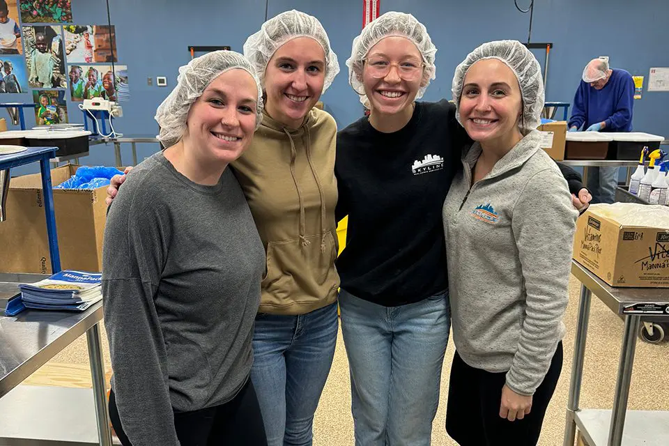 Four people wearing hairnets and casual clothes stand together smiling in a food packing facility.