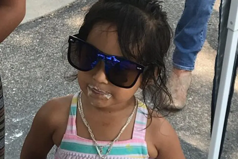 A young girl wearing large sunglasses and a striped dress stands outside with ice cream on her mouth.