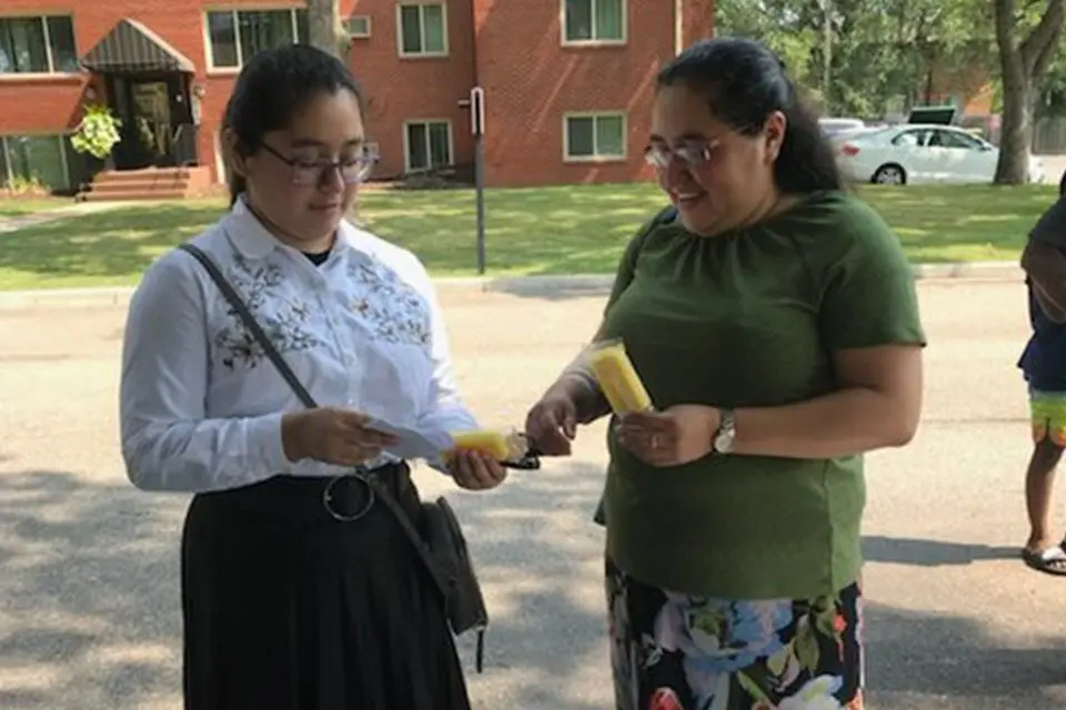 Two women standing outside, one in a white blouse and black skirt holding a popsicle, the other in a green shirt and floral skirt handing her another popsicle.