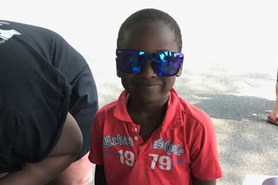 A young boy wearing oversized blue sunglasses and a red collared shirt stands outdoors, smiling at the camera.