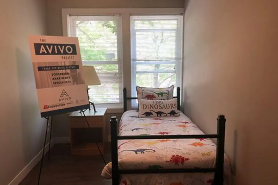 A small bedroom with a single bed featuring dinosaur bedding, a pillow reading "I Dig Dinosaurs," a wooden desk, and a sign for The Avivo Project near two windows.