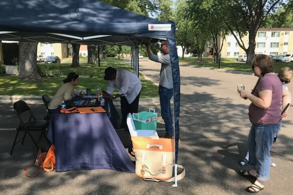 People gather under a canopy tent in a parking lot where a person sits at a table with papers and supplies, while others stand nearby and wait.