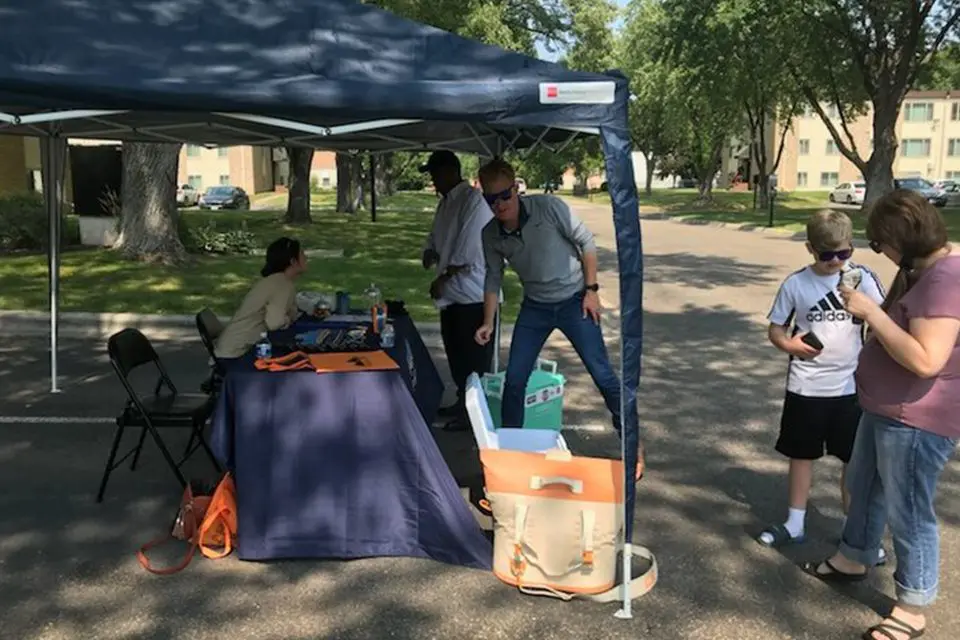 People gather under and near a pop-up canopy tent set up on a street, with a table, chairs, and bags visible. Trees and apartment buildings are in the background.
