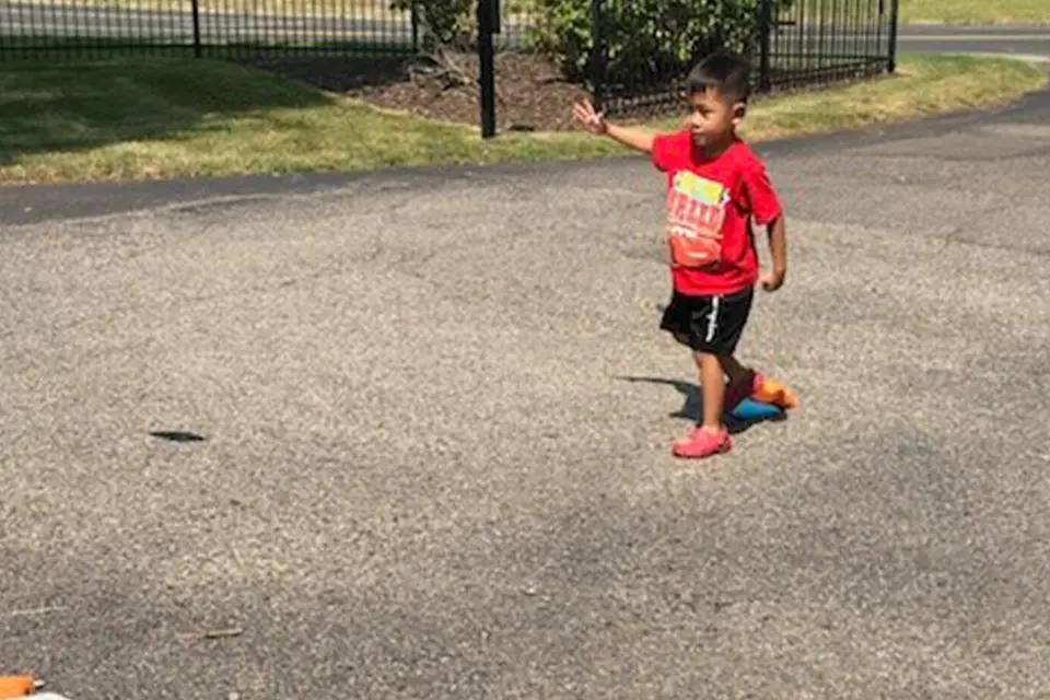 A young child in a red shirt and black shorts is walking on a paved surface outdoors, reaching out with one arm.