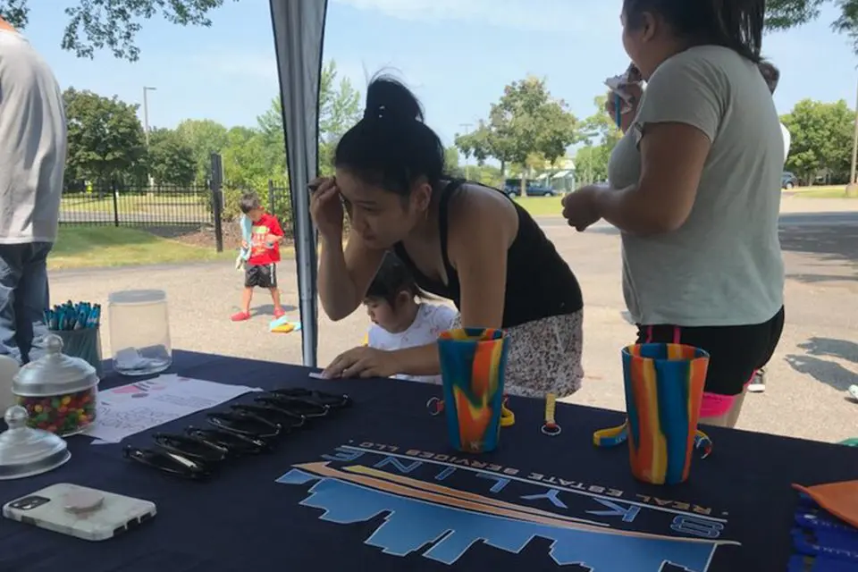 A woman leans over a table with promotional items at an outdoor event, while others stand nearby; trees and children are visible in the background.