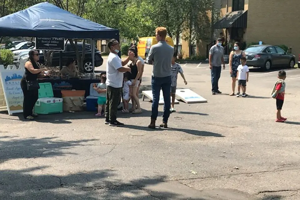 People gather near a tented booth and play cornhole in a parking lot; some are wearing masks and children are present.