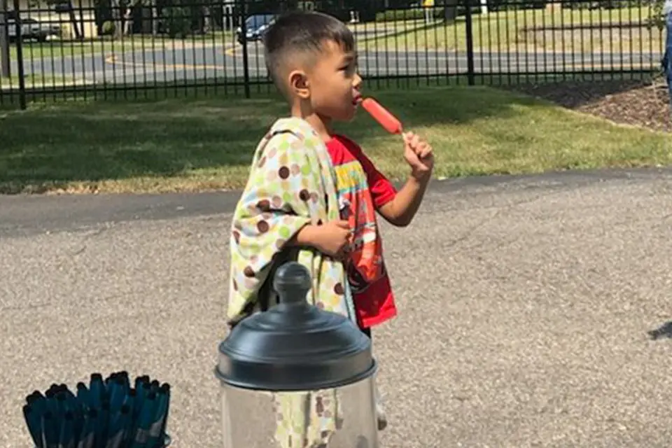 A young boy stands outside holding a blanket and eating a red popsicle, with a container and bundled items nearby on the pavement.