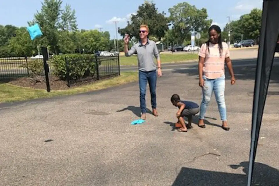 Two adults and a child play a bean bag toss game outdoors in a parking lot area on a sunny day.