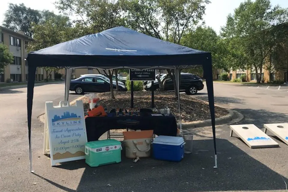 A small outdoor canopy tent with a table, coolers, and a sign advertising an ice cream party stands in a parking lot, with cornhole boards set up nearby.