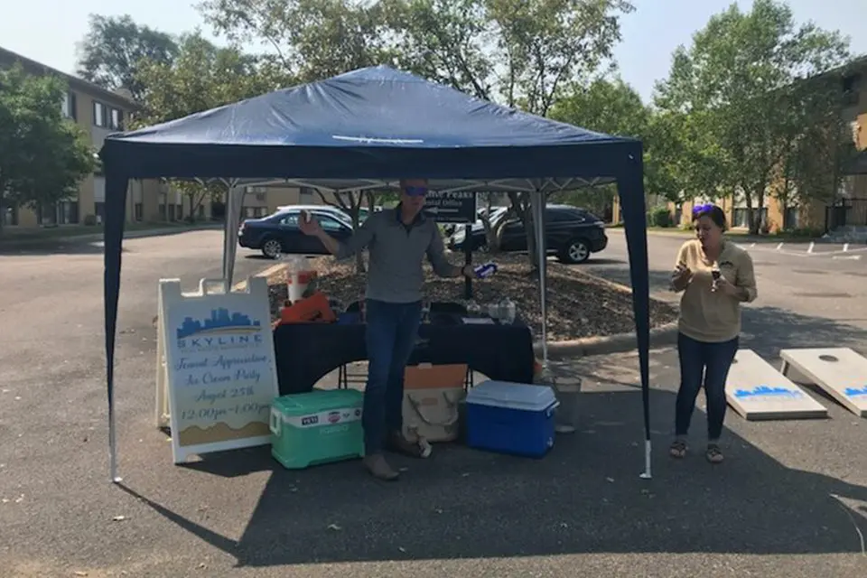 Two people stand under a blue canopy tent in a parking lot with a table, coolers, a sign, and a cornhole board nearby. Trees and parked cars are visible in the background.