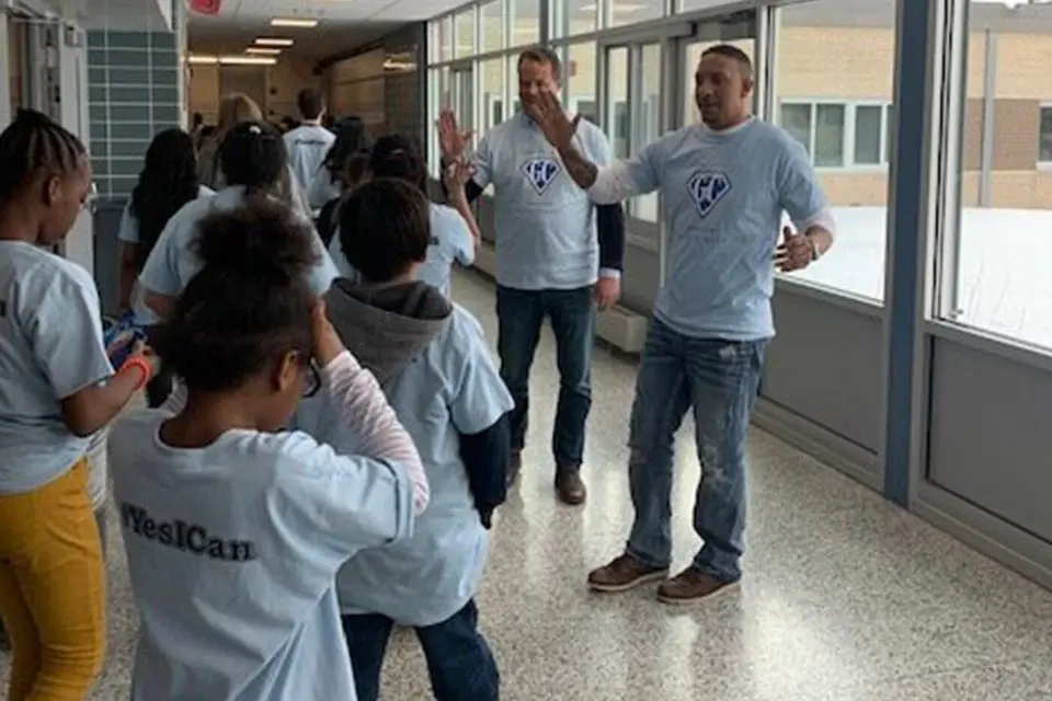 A group of people, mostly children, wearing light blue shirts walk in a school hallway. Two adults stand near the group, one gesturing with his hands.