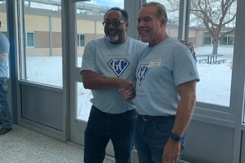 Two men wearing matching light blue t-shirts shake hands and smile while standing indoors near large windows with a snowy scene outside.