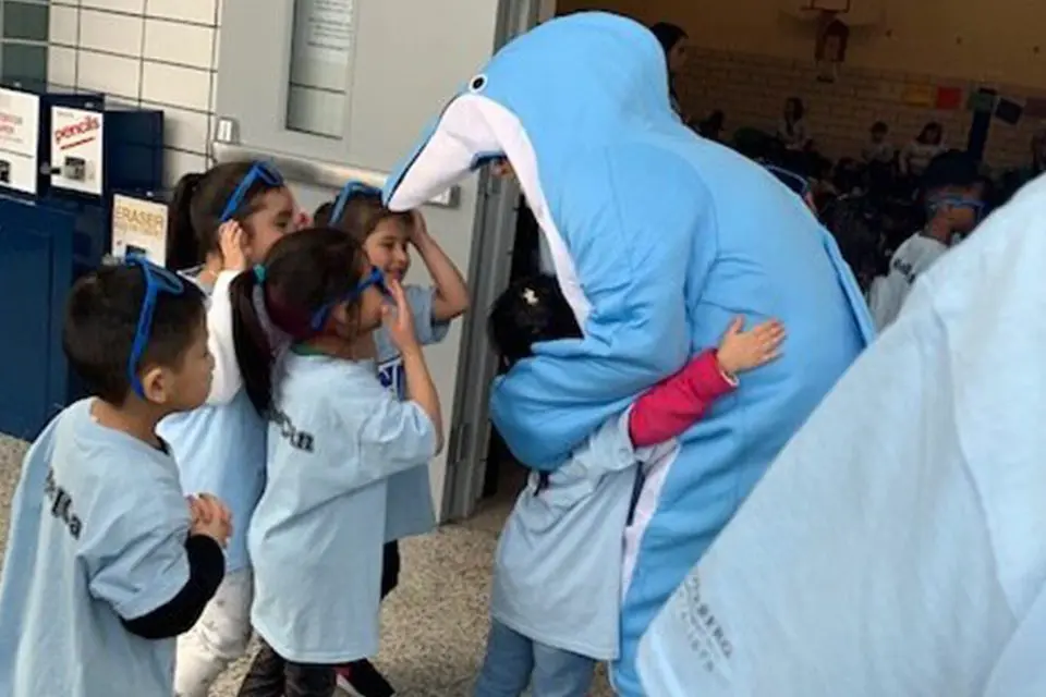 A person in a blue dolphin costume hugs and interacts with a group of young children wearing matching light blue shirts and blue sunglasses indoors.