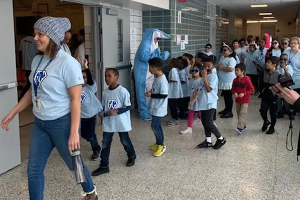 A group of children and adults wearing matching shirts walk in a school hallway; one person is dressed in a blue mascot costume.