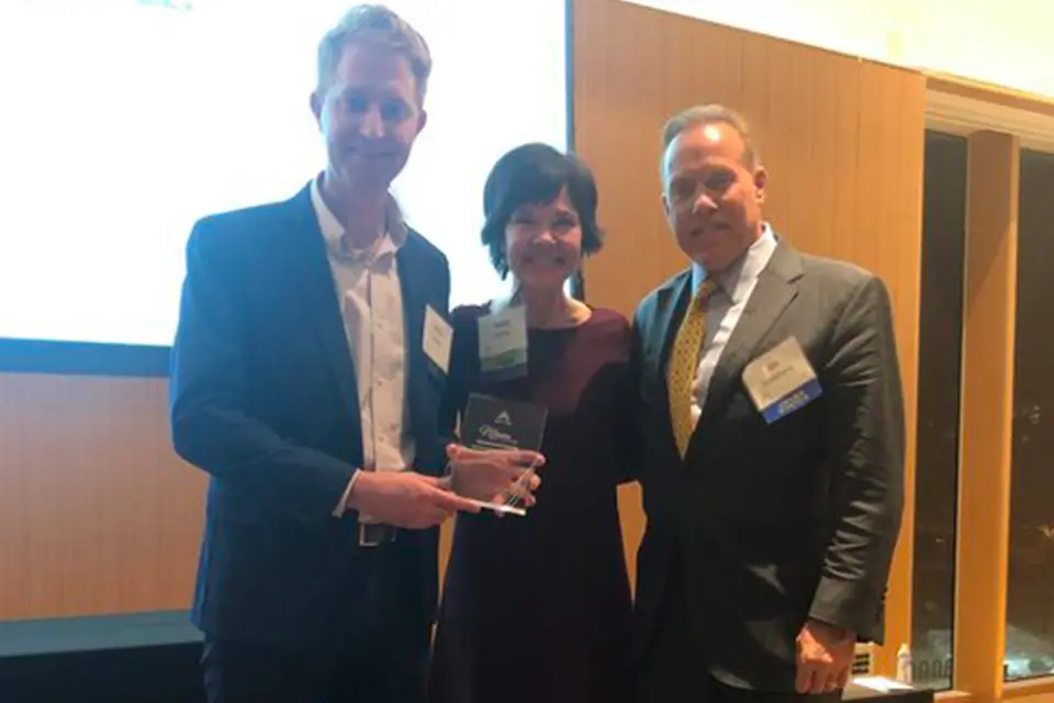 Three people in business attire stand together indoors; one holds a glass award. They are smiling and posing for a photo.
