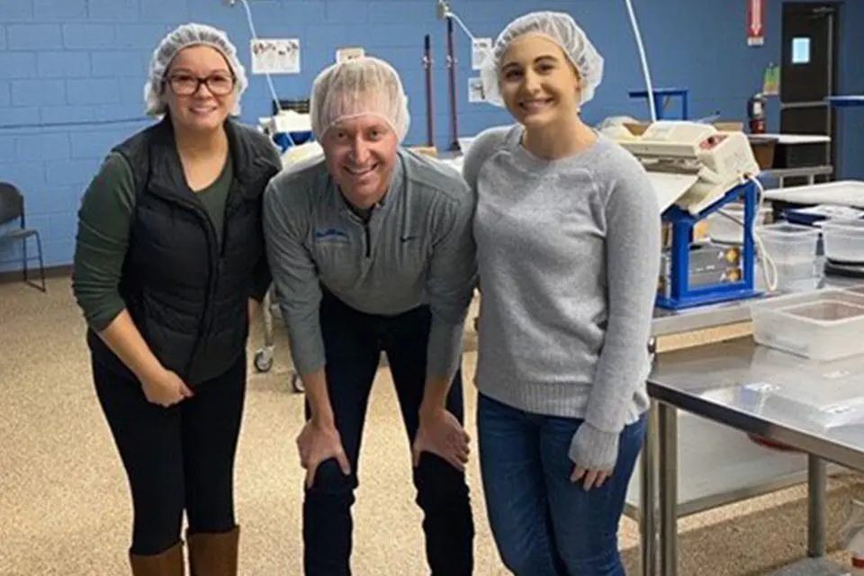 Three people wearing hair nets pose and smile in a processing or packaging facility, with equipment and tables visible in the background.