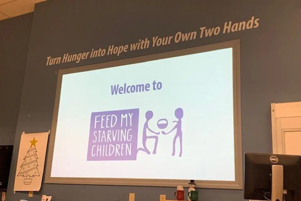 A projector screen displays "Welcome to Feed My Starving Children" with a logo, under a wall sign that reads, "Turn Hunger into Hope with Your Own Two Hands.