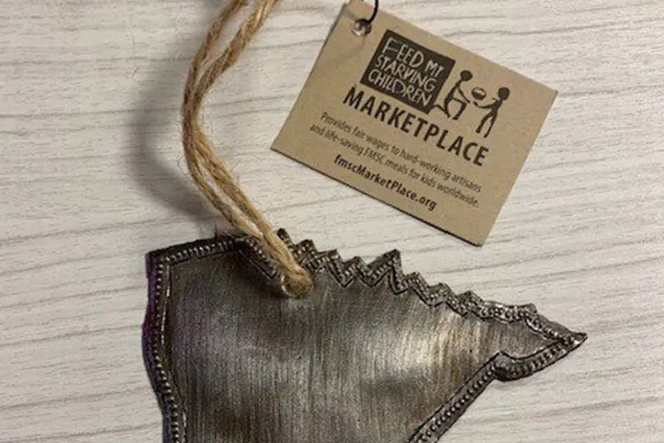 A metallic ornament shaped like the state of Minnesota with a string attached, placed on a light wood surface next to a "Feed My Starving Children Marketplace" tag.