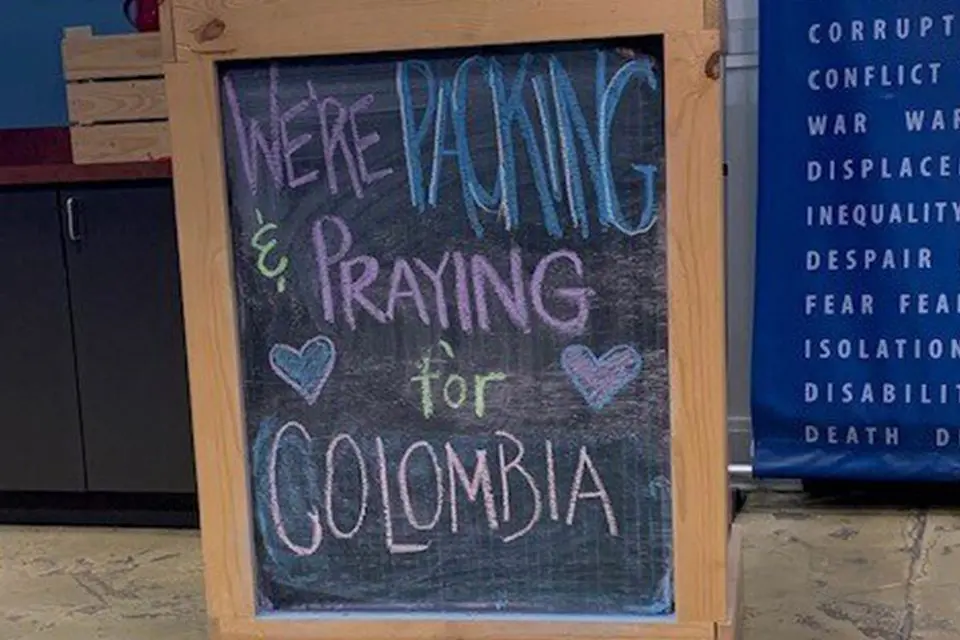 A chalkboard sign reads, "We're packing & praying for Colombia," with two drawn hearts, displayed indoors near a blue banner with white text.