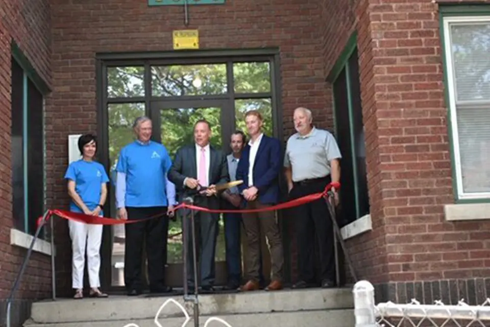 Six people stand in front of a brick building for a ribbon-cutting ceremony, with one person holding large scissors to cut the red ribbon.