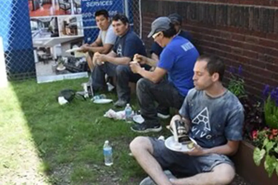 Four men sit on the grass and eat food from plates, taking a break near a brick building with a fence and some flowers nearby.