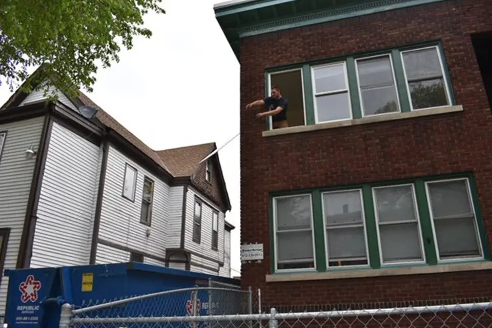 A person stands at an open window of a brick building, holding a fishing rod that extends toward a nearby house across a narrow alley.